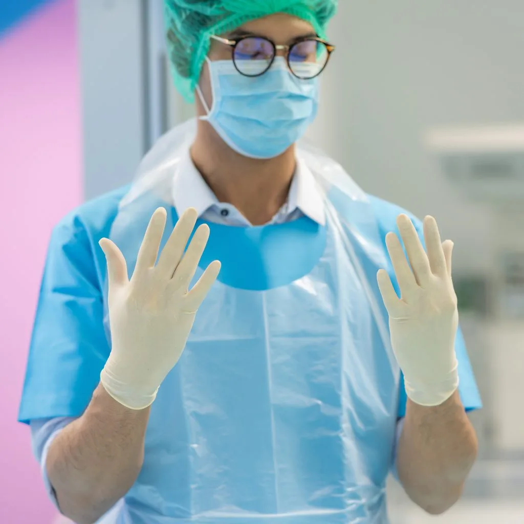 a healthcare worker wearing a blue mask, blue sterile scrubs, and white sterile gloves, holding up their hands