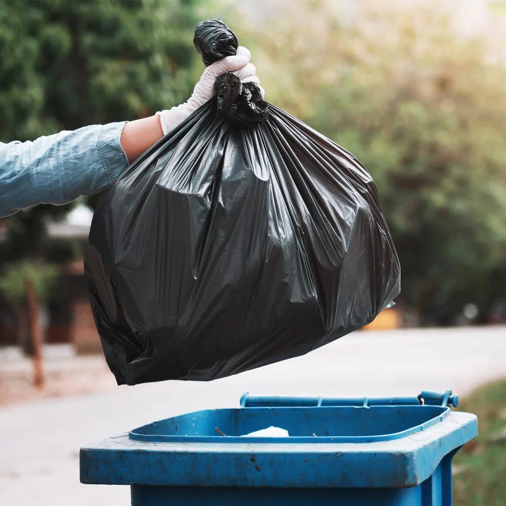 someone wearing gloves holding a full black plastic binbag over a blue wheelie bin